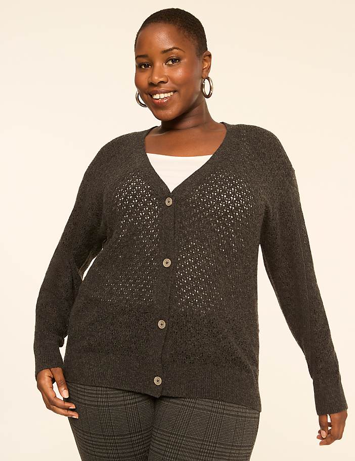 A smiling woman in a dark grey cardigan and plaid pants stands against a plain, light-colored background.