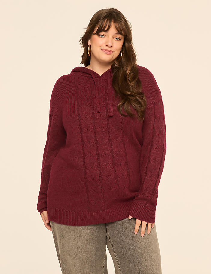 A woman in a burgundy cable-knit hoodie and gray jeans smiles against a plain, light-colored background.