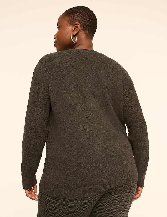 A woman in a dark gray sweater and plaid pants stands against a plain, light-colored background.