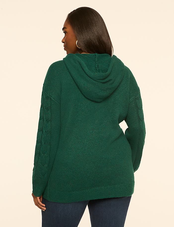 A woman in a green cable-knit hoodie stands against a plain, light-colored background.