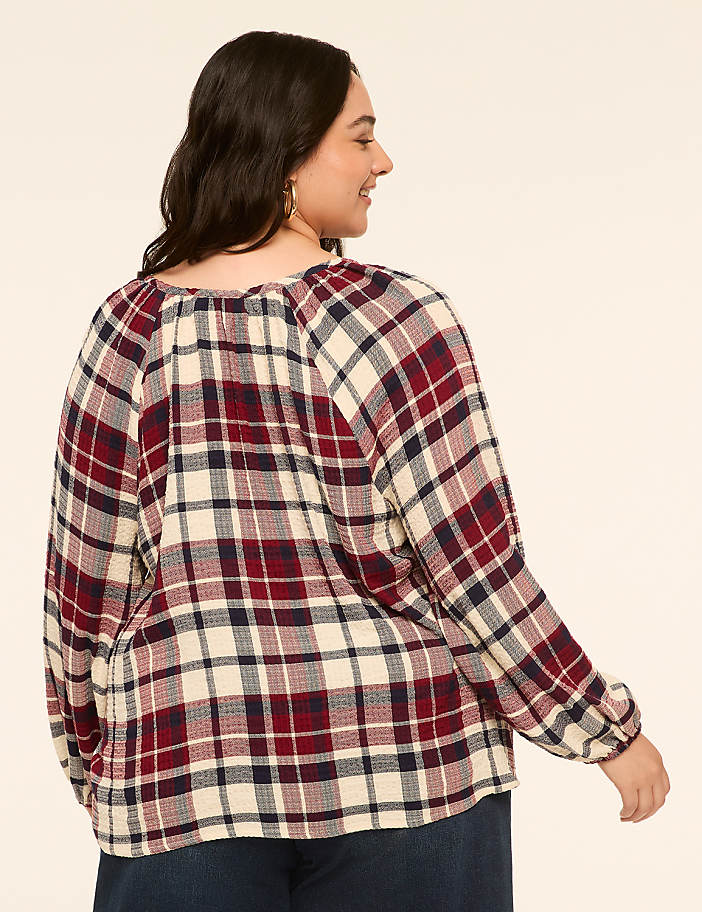 A woman in a red, white, and blue plaid shirt stands against a plain, light-colored background.