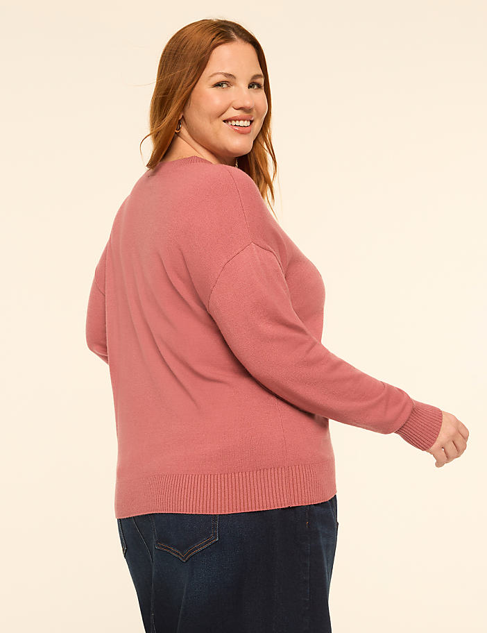 A woman in a pink sweater and dark skirt stands against a plain, light-colored background.