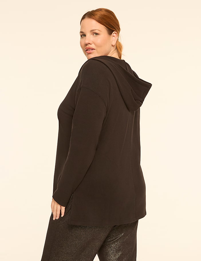 A woman in a black hooded top and sparkly pants stands against a plain, light-colored background.