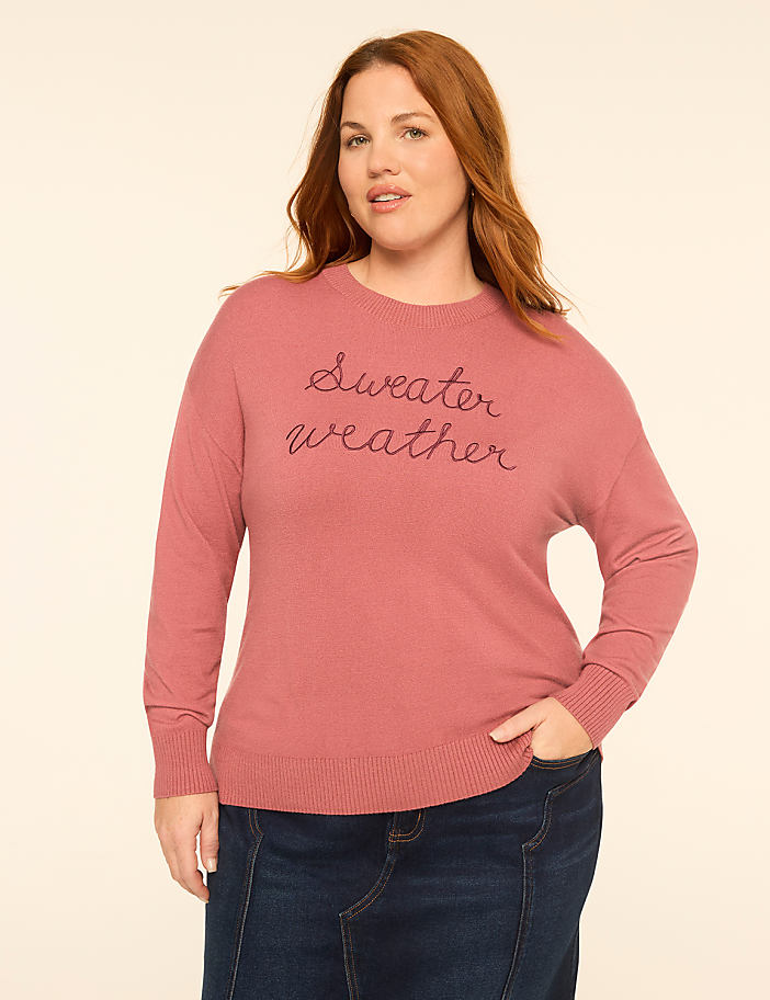 A woman with curly hair smiles in a pink "Sweater weather" sweatshirt against a plain, light-colored background.