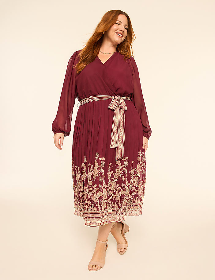 A smiling woman in a burgundy wrap dress with a patterned hem stands against a plain, light-colored background.