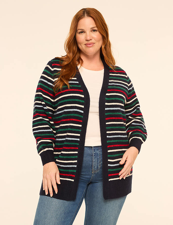 A woman with red hair stands in the foreground wearing a striped cardigan over a white top and blue jeans, against a plain, light-colored background.