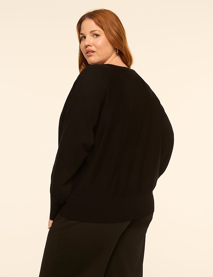 A woman with red hair wearing a black sweater and pants stands against a plain, light-colored background.