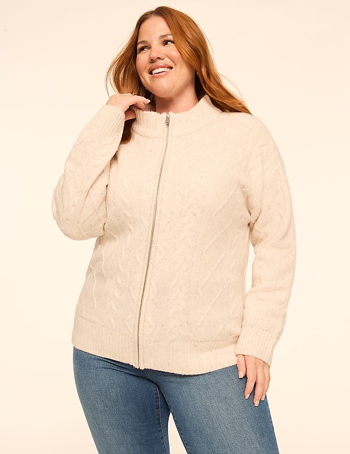 A smiling woman in a cream cable-knit zip-up sweater and blue jeans stands against a plain, light beige background.