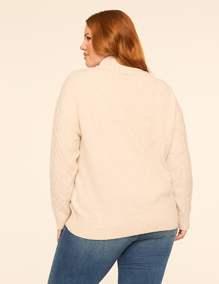 A woman with red hair wearing a cream cable-knit sweater and blue jeans stands against a plain, light-colored background.