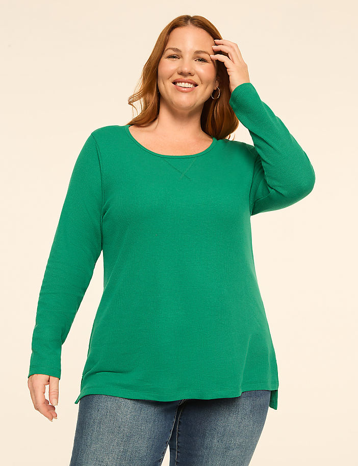 A smiling woman with auburn hair in a green waffle-knit top and blue jeans stands against a plain, light-colored background.