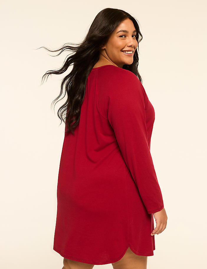 A smiling woman in a red dress stands against a plain, light-colored background.