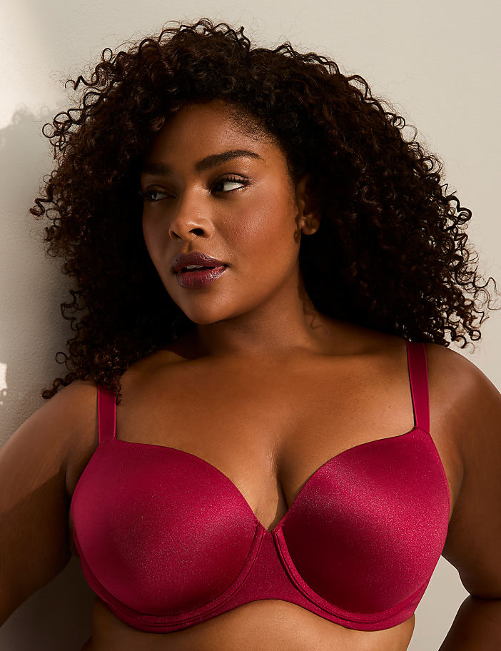 A woman with curly dark hair and a maroon bra is leaning against a white wall.