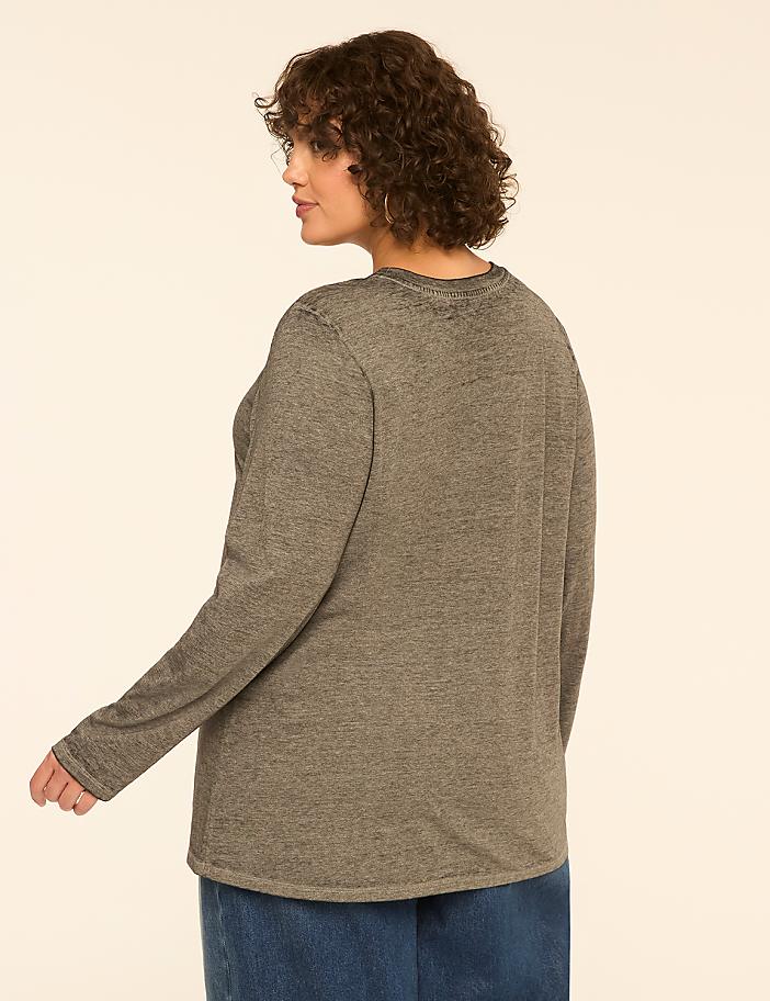 A woman with curly brown hair, wearing a gray long-sleeved shirt and a blue skirt, stands against a plain, light-colored background.