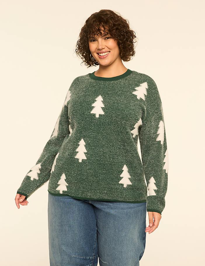 A smiling woman in a fuzzy green sweater with white Christmas trees stands against a plain, light-colored background.