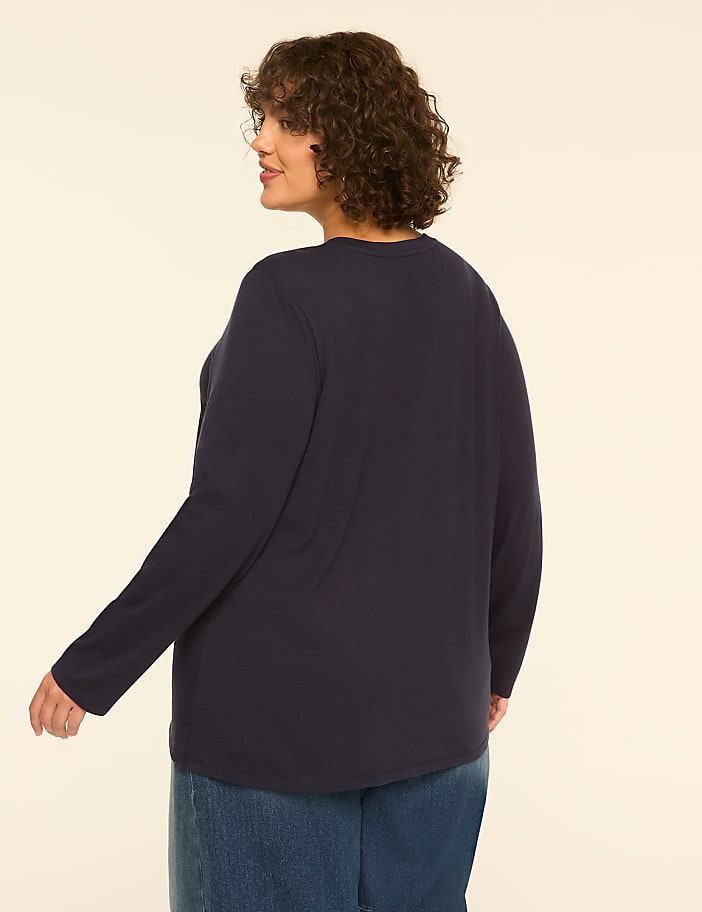 A woman in a navy long-sleeved shirt and blue jeans stands against a plain, light-colored background.