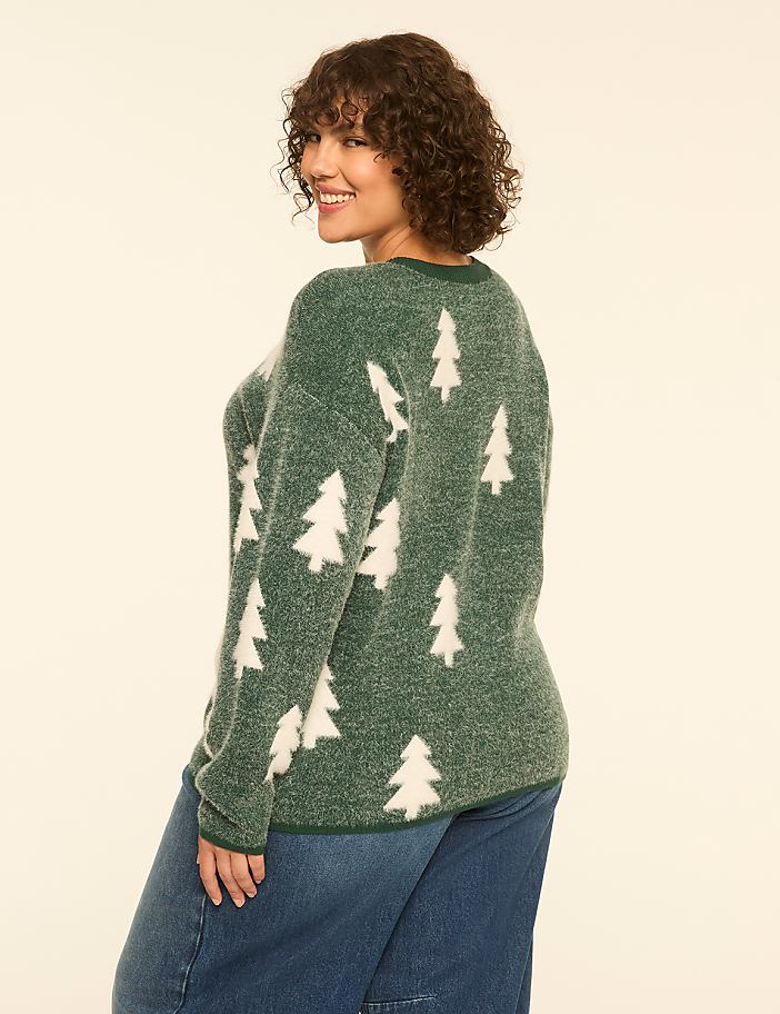 A woman in a green, fuzzy sweater with white Christmas trees stands against a plain, light-colored background.