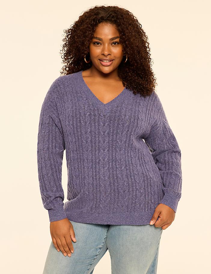 A woman with curly hair wearing a purple cable-knit V-neck sweater and light blue jeans stands against a plain, light-colored background.
