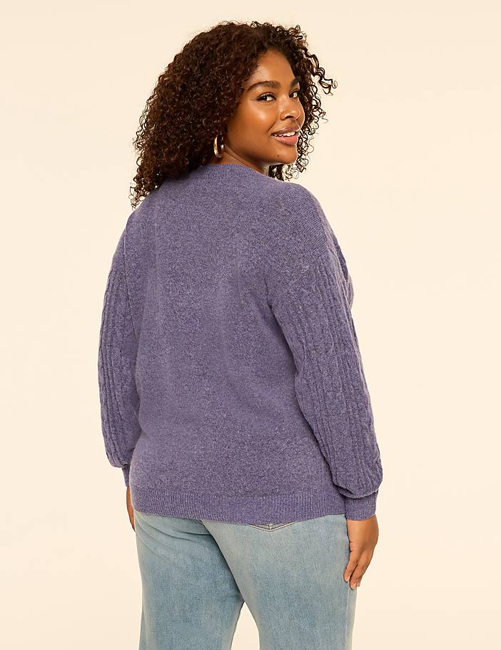 A woman with curly hair wearing a purple sweater and light blue jeans stands against a plain, light-colored background.