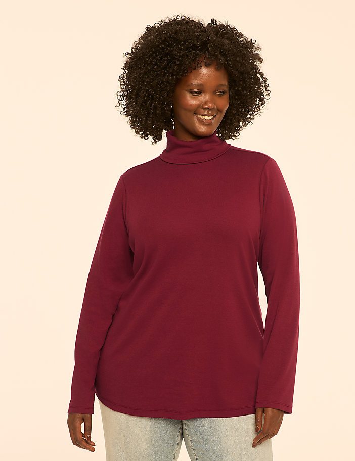 A smiling woman with curly dark hair wears a maroon turtleneck and light blue jeans against a plain cream background.