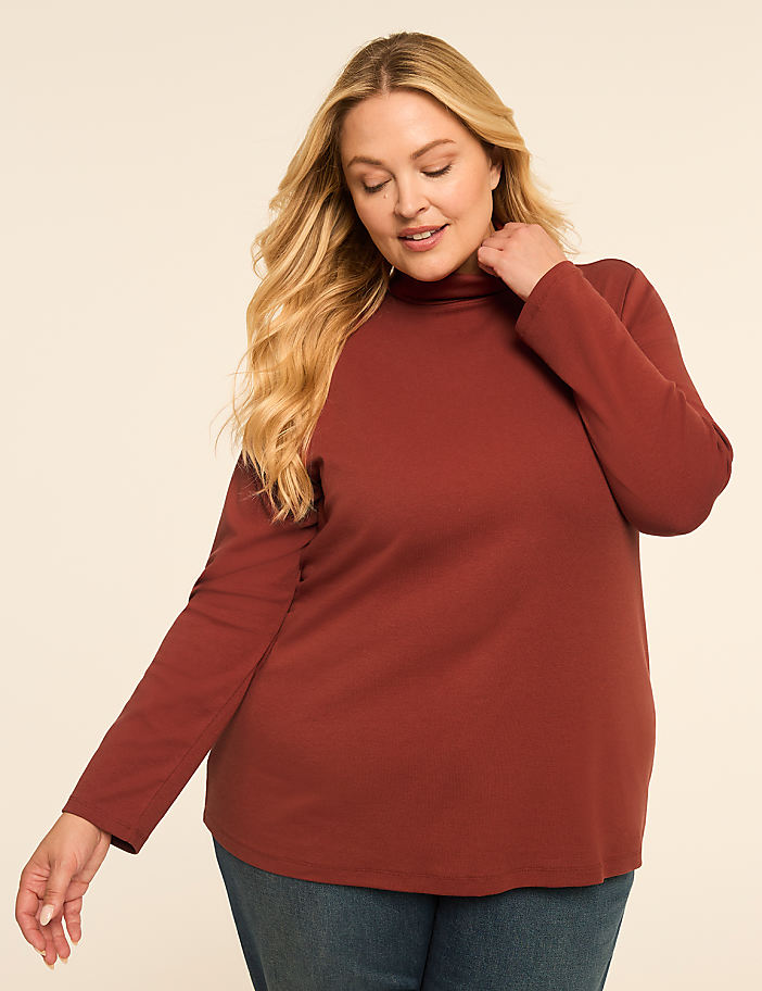 A woman in a rust-colored turtleneck and jeans stands against a plain, light-colored background.