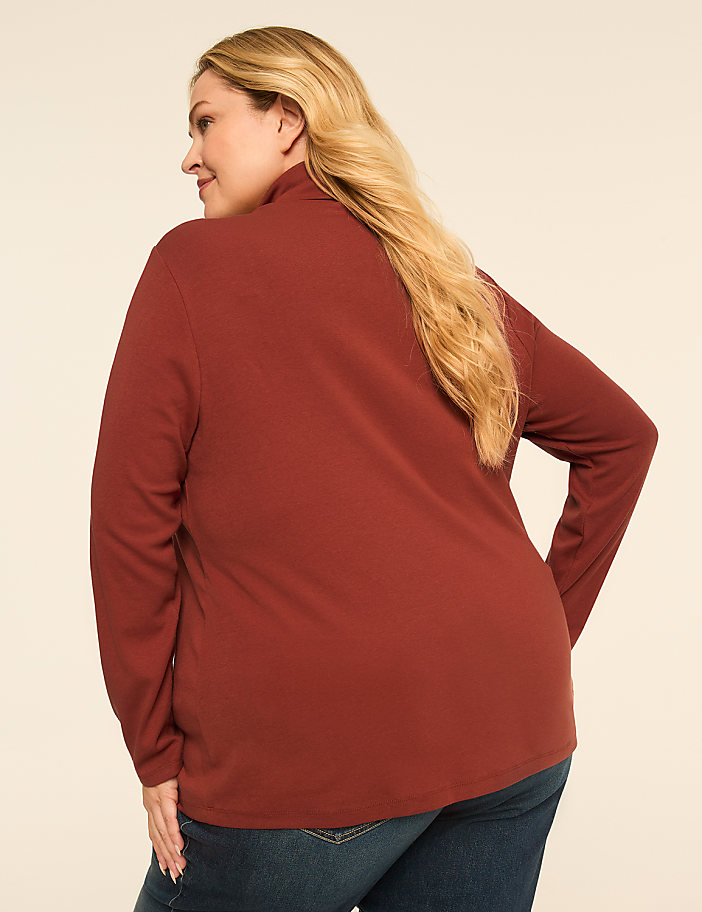 A woman in a rust-colored turtleneck and blue jeans stands against a plain, light-colored background.