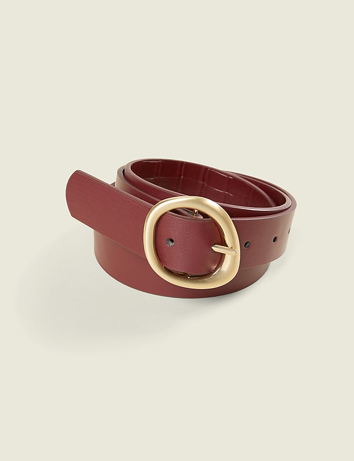 A rolled burgundy leather belt with a gold oval buckle sits against a plain, light beige background.