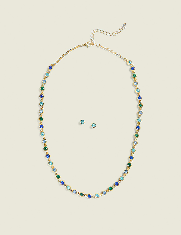 A gold necklace with blue and green gemstones and matching stud earrings are centered on a plain, light beige background.