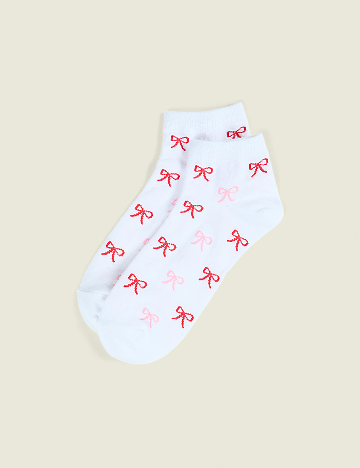 A pair of white socks with small red bows are laid flat against a plain, light beige background.
