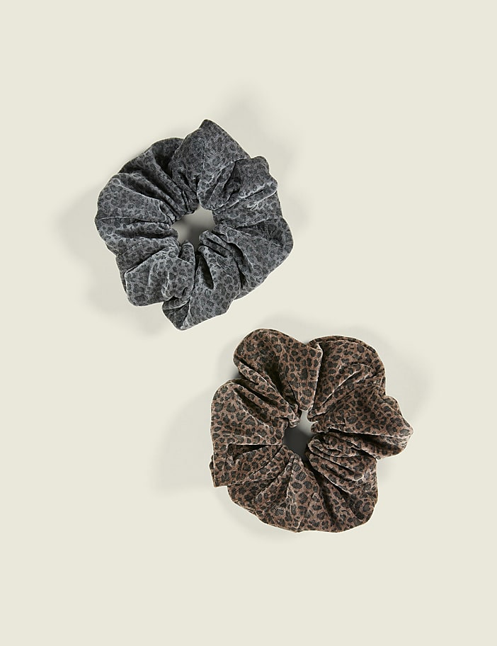 Two leopard-print scrunchies, one grey and one brown, lie on a plain off-white background.