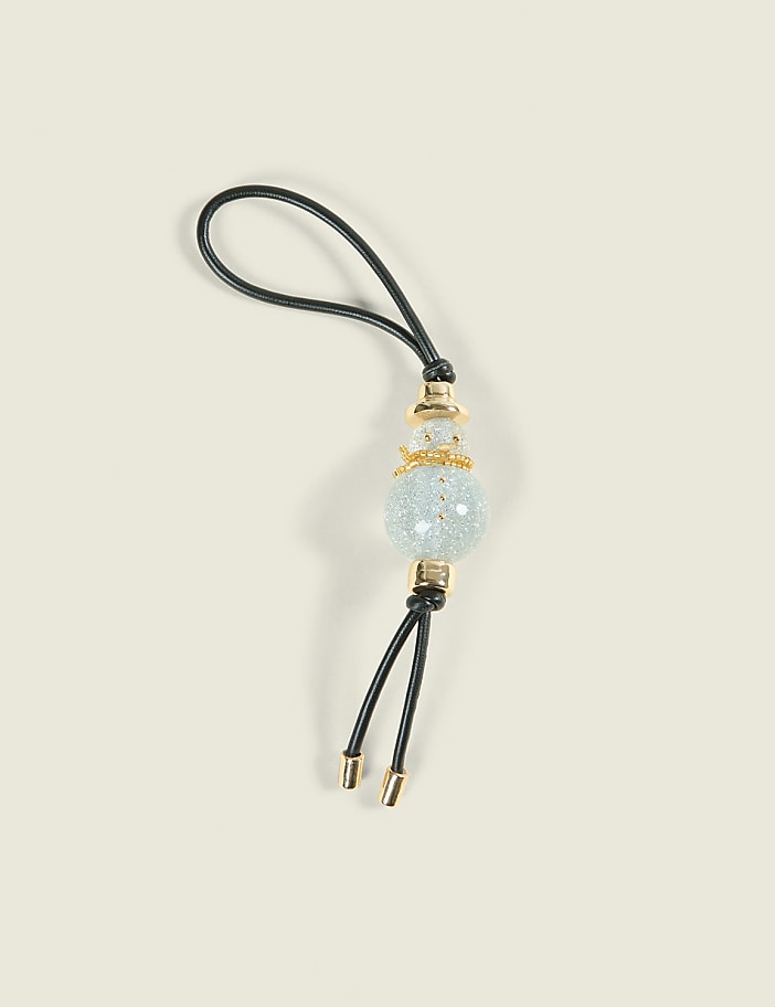 A decorative snowman charm with a glittery blue bead and gold accents hangs from a black cord against a plain, light beige background.