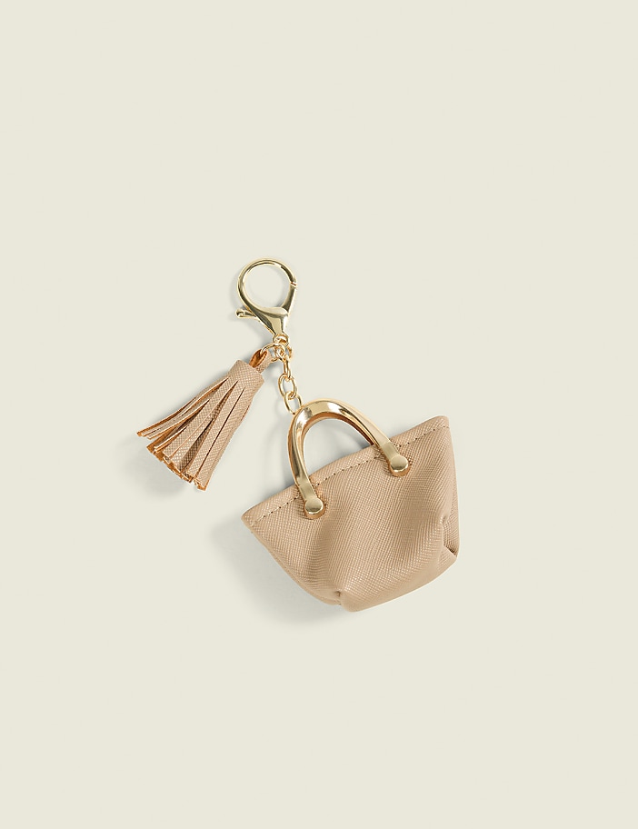 A beige handbag keychain with a gold tassel rests on a plain, light-colored background.