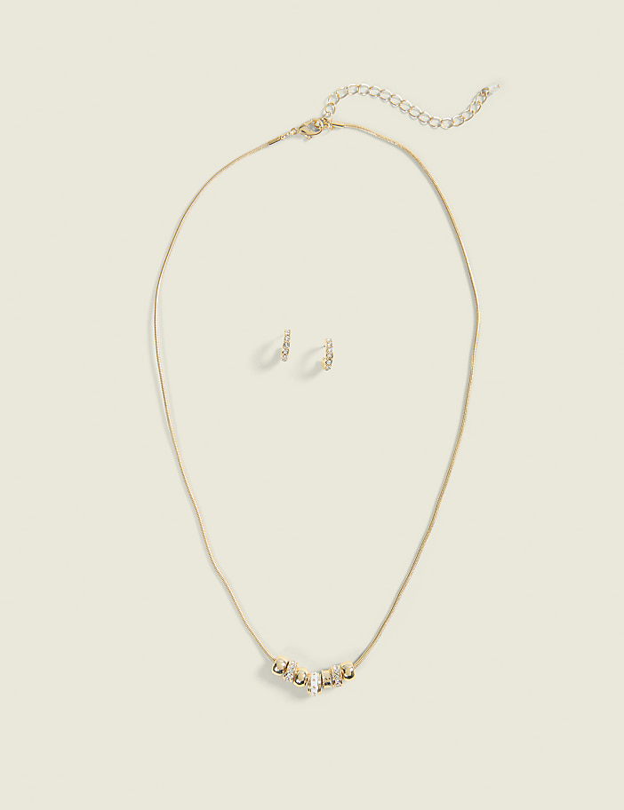 A gold necklace with a crystal-embellished pendant and matching stud earrings are laid out on a plain, light-beige background.