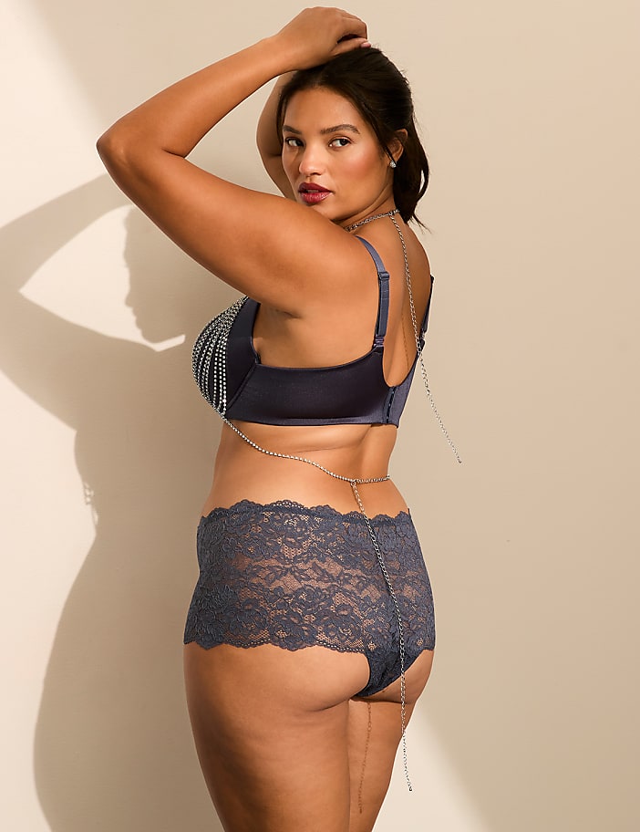 A woman in blue lingerie and a body chain poses against a plain, light-colored wall.