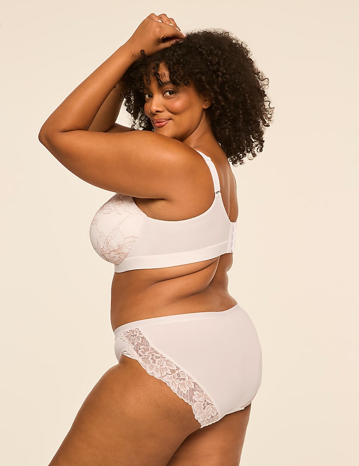 A woman with curly hair wearing white lace lingerie poses against a plain, light-colored background.