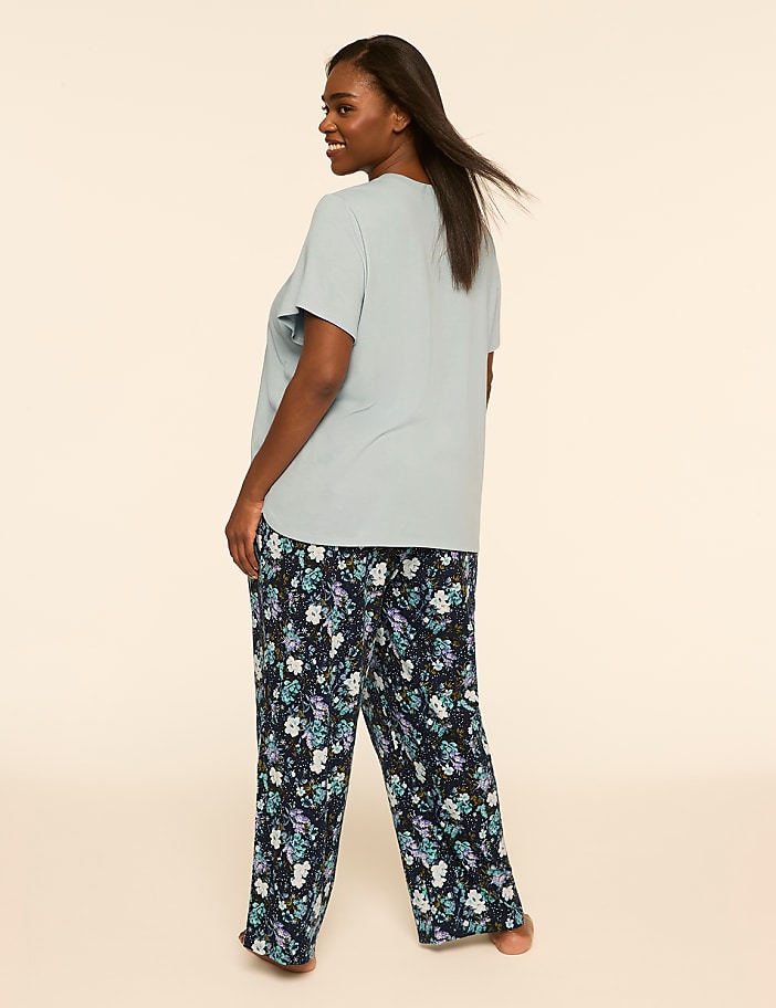 A woman wearing a light blue t-shirt and floral print pajama pants stands against a plain, neutral background.