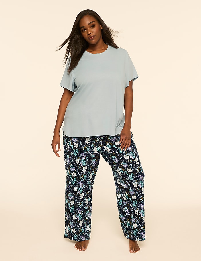 A woman wearing a light blue t-shirt and floral print pants stands against a plain, neutral background.