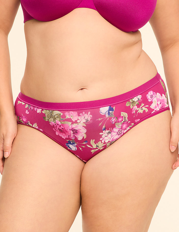 A woman wears a magenta bra and floral print panties against a plain, light-colored background.