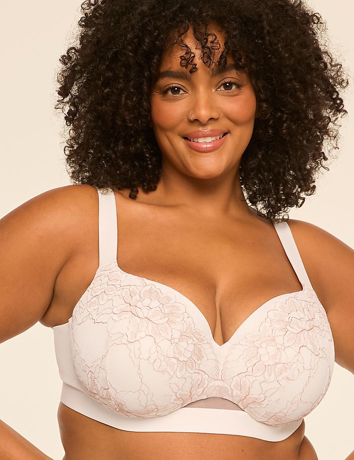 A smiling woman with curly dark hair wearing a white lace bra poses against a plain, light-colored background.