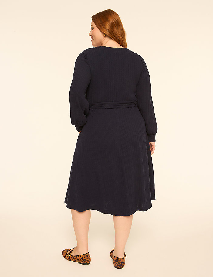 A woman with red hair wearing a navy blue long-sleeved dress and leopard print flats stands against a plain off-white background.