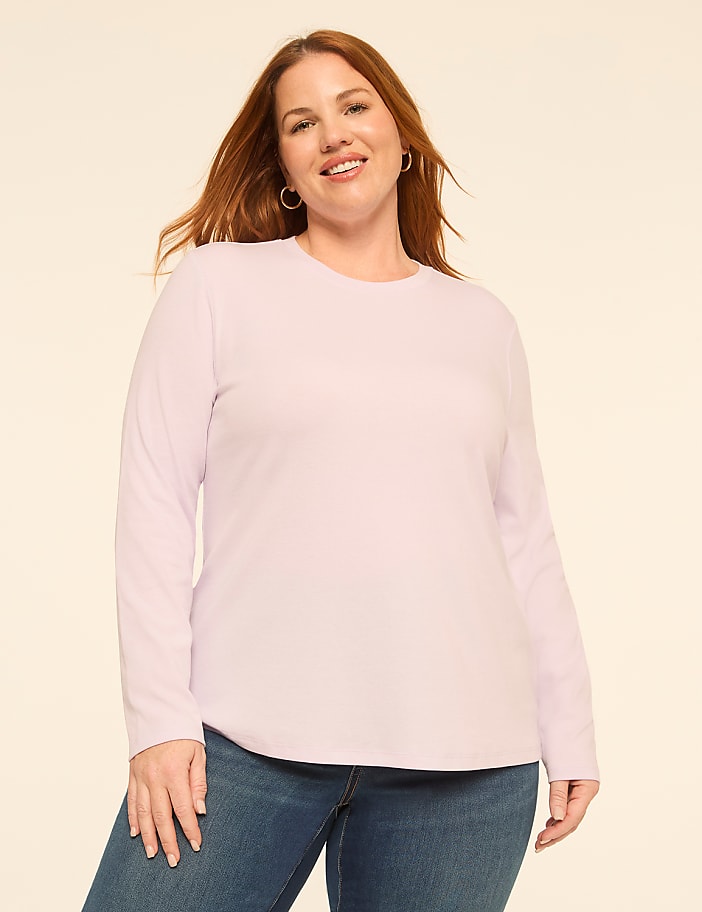 A smiling woman with red hair wearing a light purple long-sleeved shirt and blue jeans stands against a plain, off-white background.