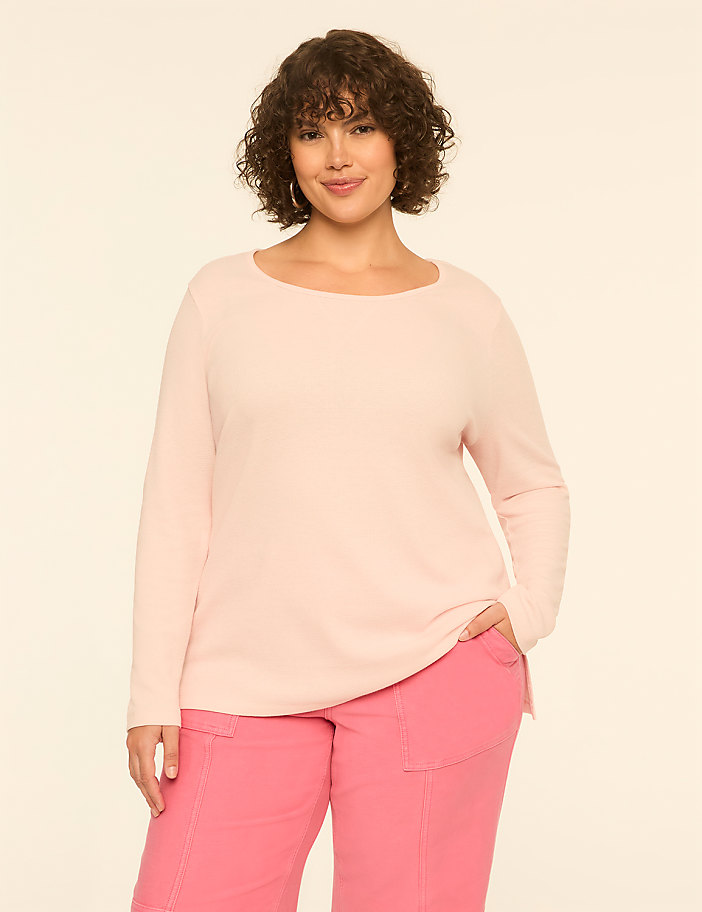 A woman with curly hair wearing a light pink long-sleeved top and bright pink pants stands against a plain, off-white background.