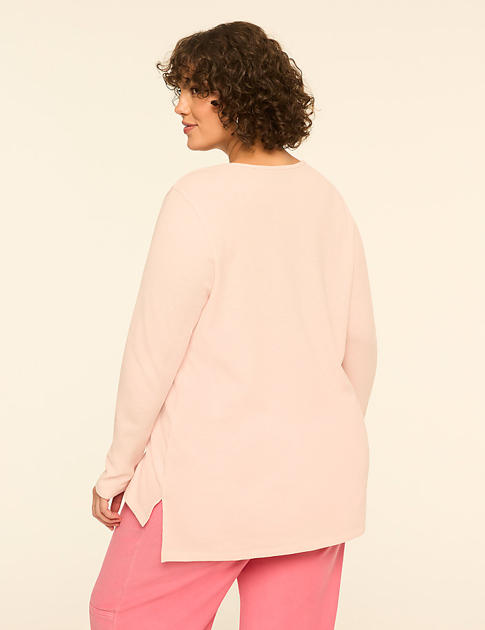 A woman with curly brown hair wearing a light pink long-sleeved top and bright pink pants stands against a plain, off-white background.