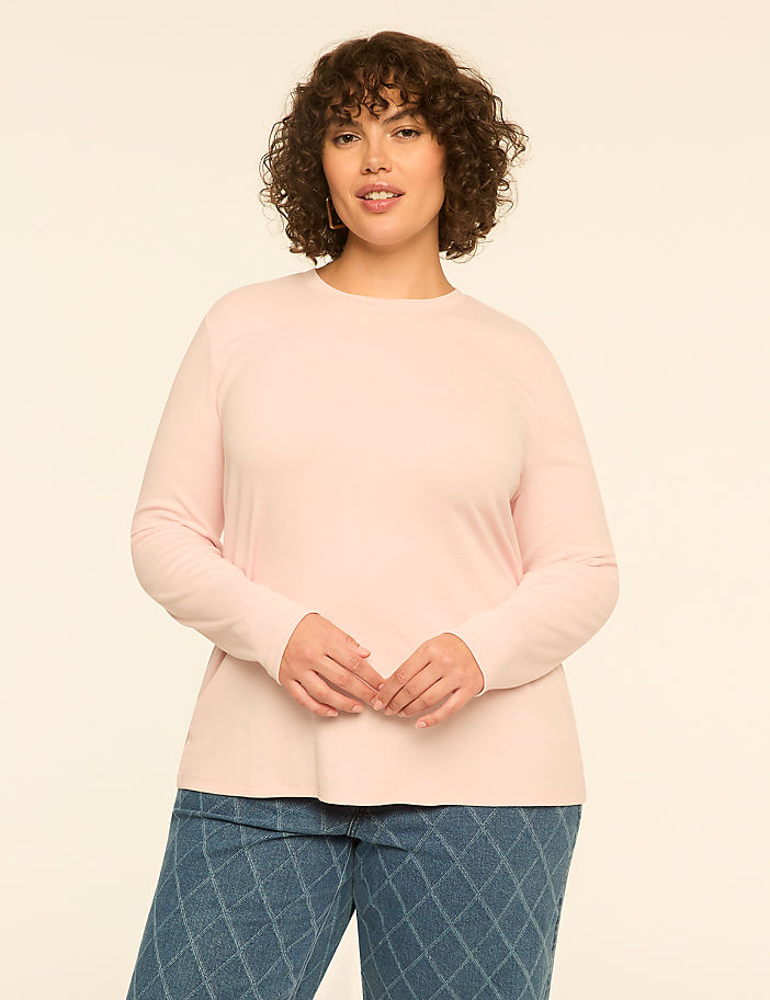 A woman with curly brown hair wearing a light pink long-sleeved top and patterned blue jeans stands against a plain off-white background.