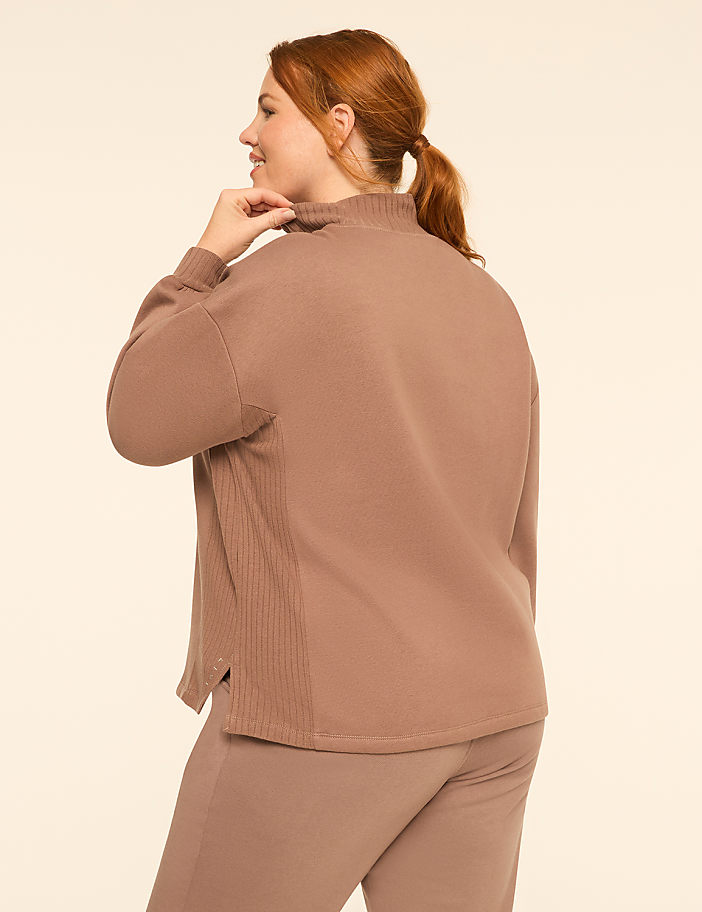 A woman with red hair tied in a ponytail wears a brown lounge set against a plain, light-colored background.