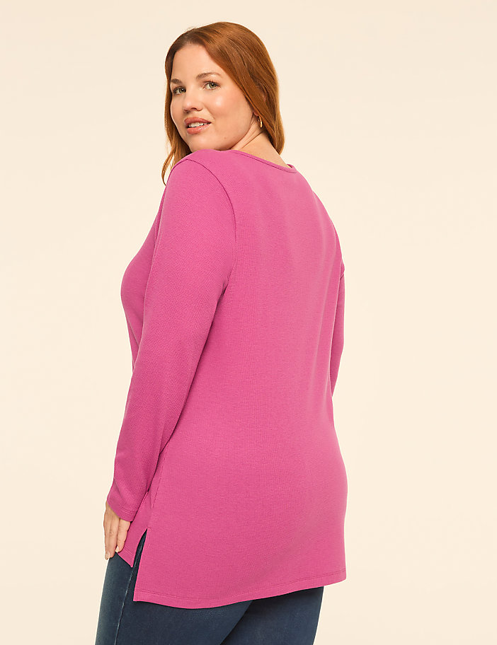 A woman with red hair wearing a long-sleeved pink top and blue jeans stands against a plain, light-colored background.