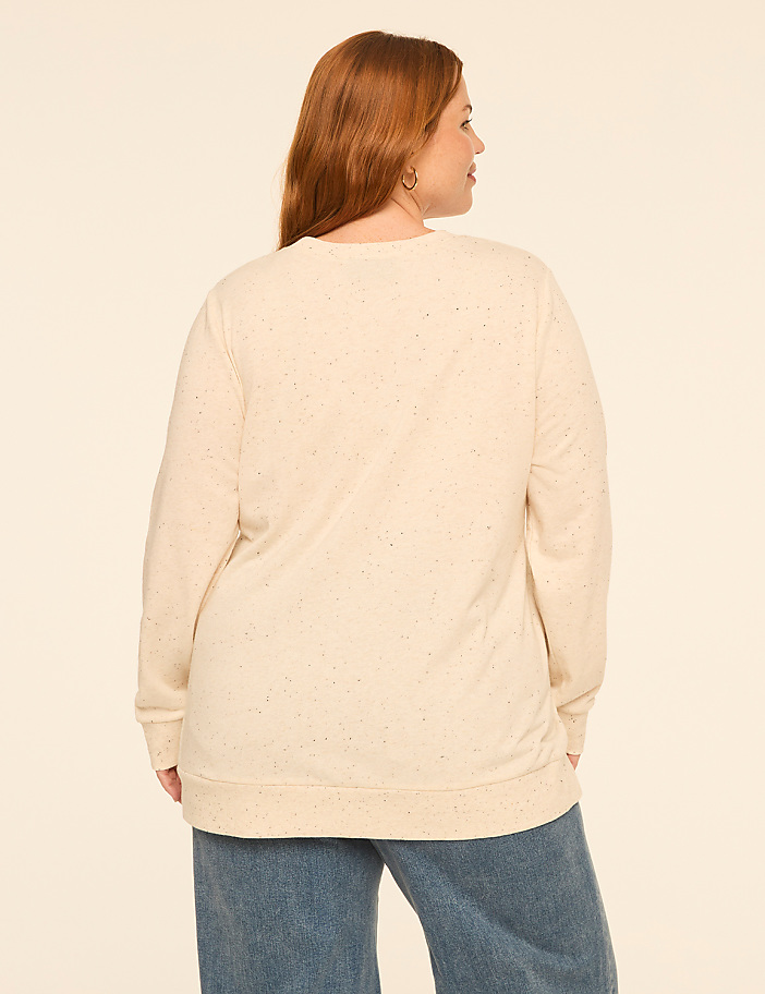 A woman with red hair wearing a cream-colored speckled sweatshirt and blue jeans stands against a plain, light-colored background.