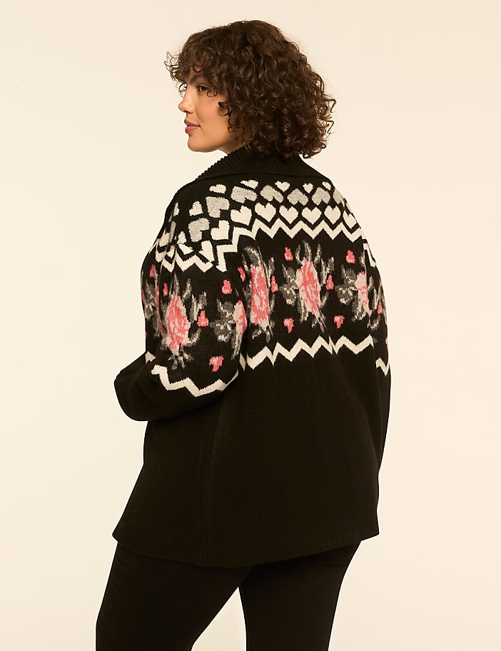 A woman with curly brown hair wearing a patterned black cardigan and black pants stands against a plain white background.