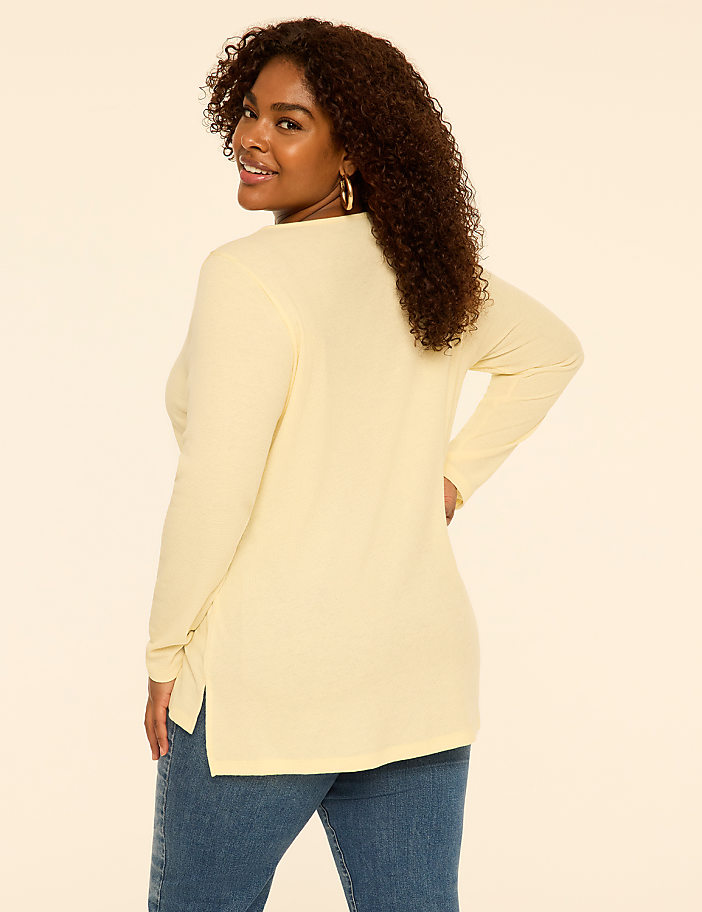 A woman with curly hair wearing a pale yellow long-sleeved top and blue jeans stands against a plain, light-colored background.