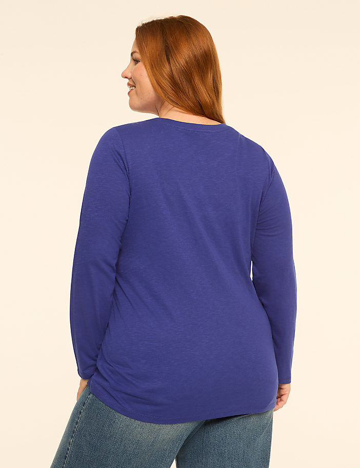 A woman with red hair wearing a blue long-sleeved shirt and jeans stands against a plain, light-colored background.