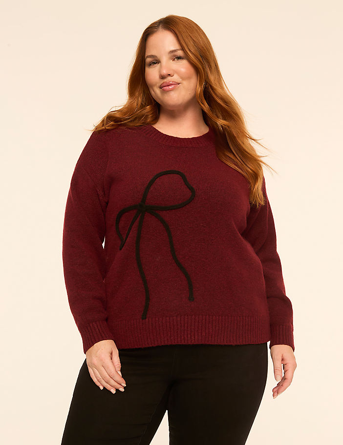 A woman with long red hair wearing a burgundy sweater with a black bow design and black pants stands against a plain off-white background.